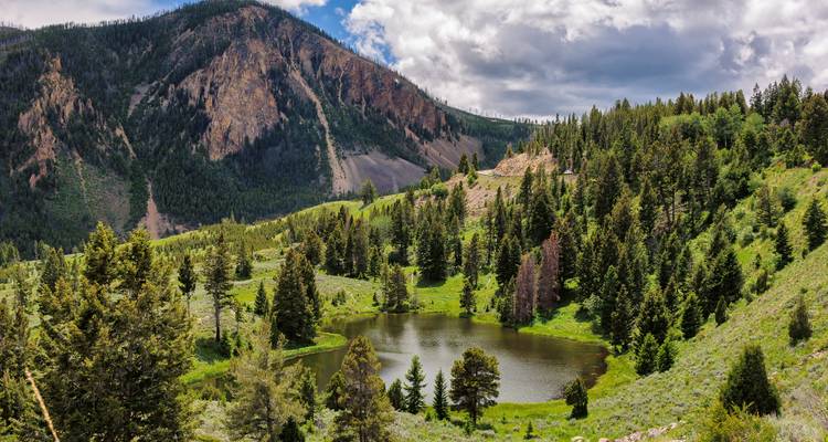 Lago alpino rodeado de denso bosque de coníferas con montañas escarpadas al fondo bajo un cielo azul parcialmente nublado.