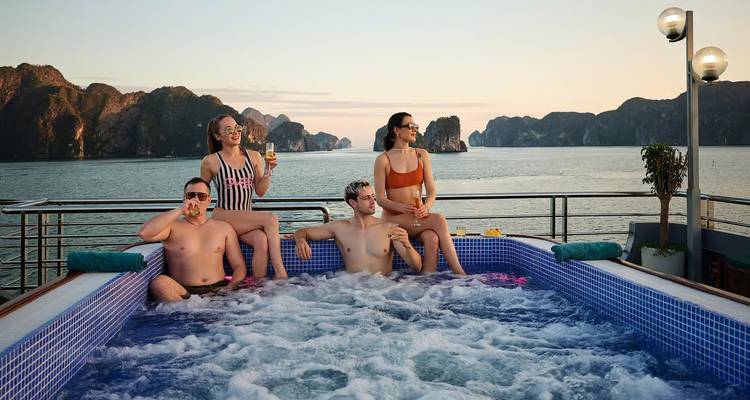 Four friends relax in a bubbling rooftop Jacuzzi on a cruise ship with Halong Bay karst islands behind them at sunset.