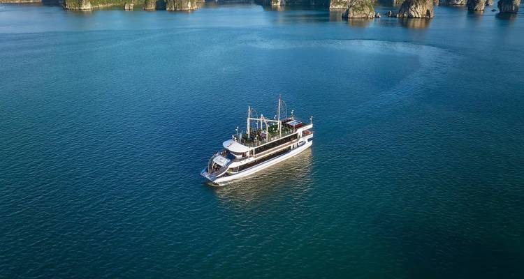 Aerial view of a white day-cruise boat alone on the emerald waters of Halong Bay surrounded by limestone islands.