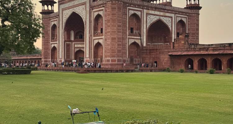 Imposante zandstenen ingangspoort van het Taj Mahal complex kijkt uit op een verzorgd gazon vol bezoekers.