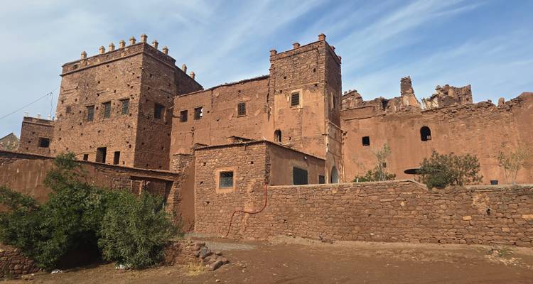 Ruinas de kasbah de color tierra con torres cuadradas y almenas ornamentadas bajo un cielo azul, situadas en una ladera rocosa.