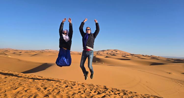 Dos viajeros saltan alegremente hacia el aire en la cima de una duna de arena dorada con un desierto sin fin detrás de ellos.