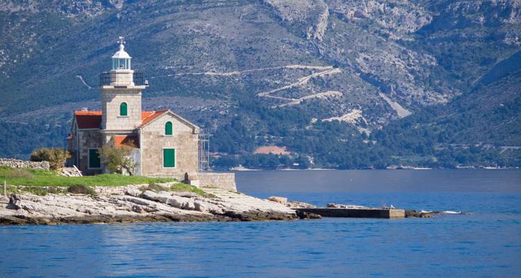 Phare en pierre avec toit en tuiles rouges se dressant sur un rivage rocheux croate, dominé par des montagnes boisées.