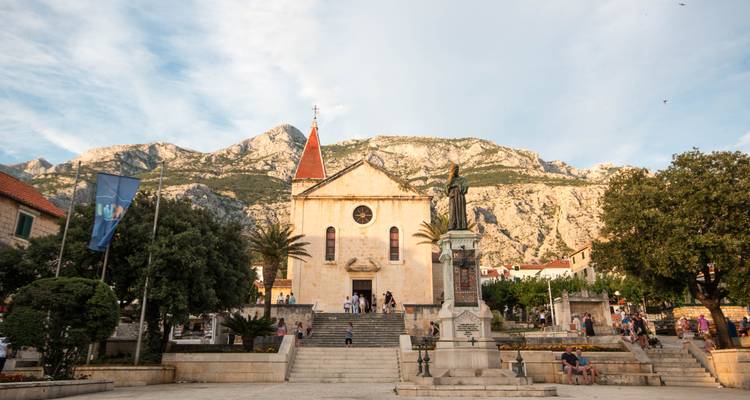 Place de la ville à Makarska avec église, chaîne de montagnes en arrière-plan et lumière du soir tandis que les habitants flânent sur les marches.