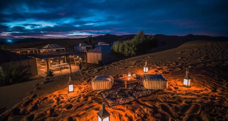 Campamento desértico con ambiente mágico al atardecer con asientos iluminados por linternas en las dunas de arena contra un cielo azul púrpura espectacular.