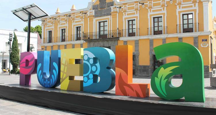 Buntes dreidimensionales "PUEBLA"-Schild vor einem gelben Kolonialgebäude.