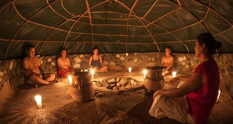 Menschen, die um Kerzen herum in einer traditionellen Temazcal-Schwitzhütte aus Stein und Holz sitzen.