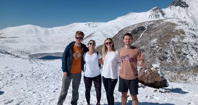 Vier Freunde stehen in schneebedecktem Gelände mit dem Krater und dem gefrorenen See des Nevado de Toluca im Hintergrund.