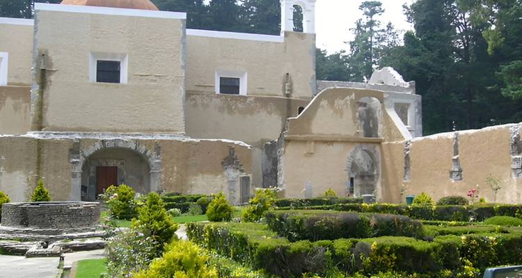 Ein historischer beiger Klosterkomplex mit gepflegten Heckengartenanlagen in einem Kiefernwald.