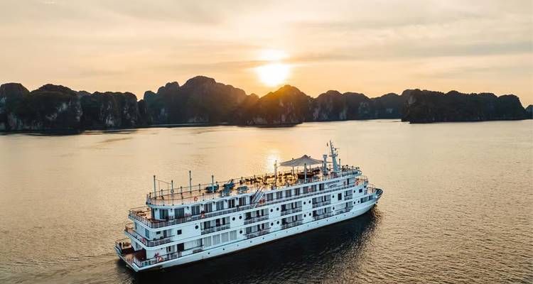 A white cruise ship sails through tranquil waters at sunset with limestone karsts silhouetted behind