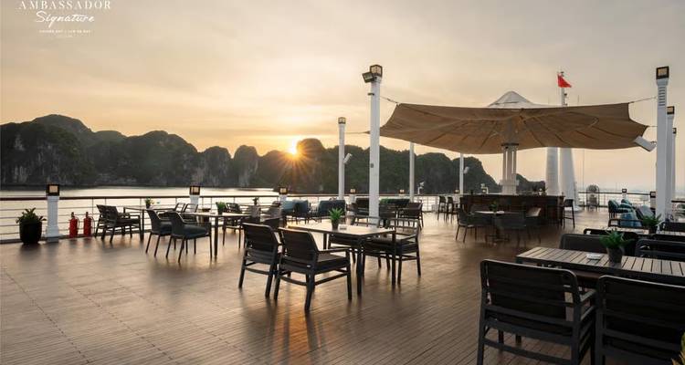 Open-air dining deck with tables and chairs overlooking the bay as the sun drops behind rugged outcrops