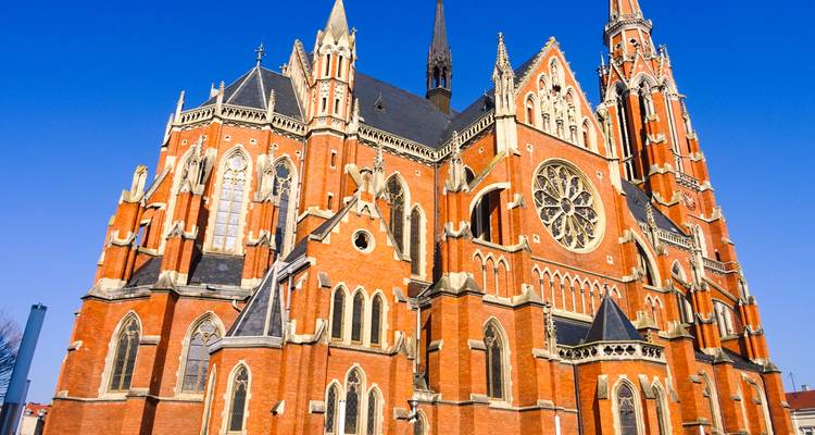 Striking red-brick neo-gothic cathedral with ornate spires and rose window set against a clear blue sky in Osijek.