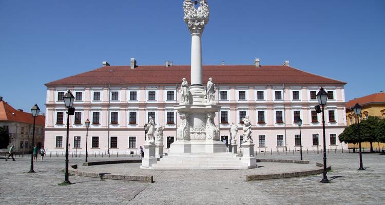 Open cobblestone square featuring a white Baroque plague column surrounded by classical buildings under a cloudless sky.