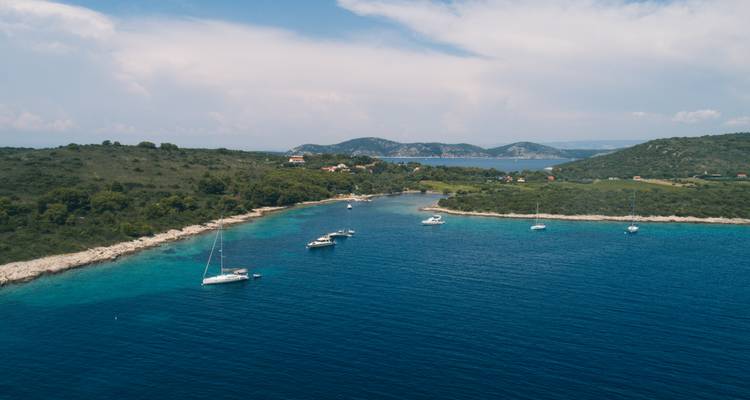 Luftaufnahme einer ruhigen Bucht mit Segelbooten, die in der Nähe einer bewaldeten kroatischen Insel vor Anker liegen, und türkisfarbenem Wasser.