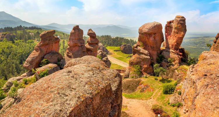 Formaciones de arenisca roja dramáticas que se elevan sobre un valle verde con montañas lejanas y brumosas.
