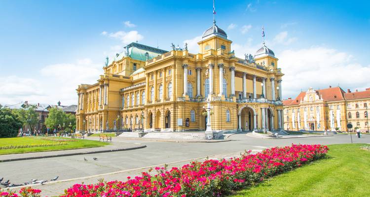 Historic yellow Croatian National Theatre in Zagreb with manicured flower beds and blue sky.