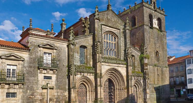 Massieve stenen gevel van de Kathedraal van Porto met ingewikkelde gotische bogen en roosvenster tegen een felblauwe hemel.