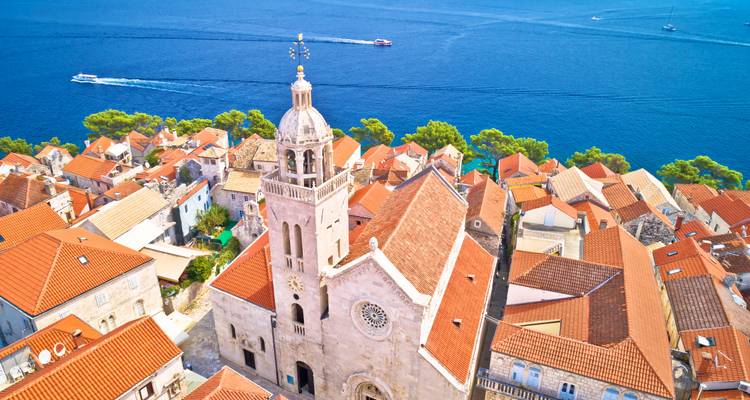 Aerial view of a stone church tower rising above orange roofs with deep blue Adriatic waters and speedboats beyond.