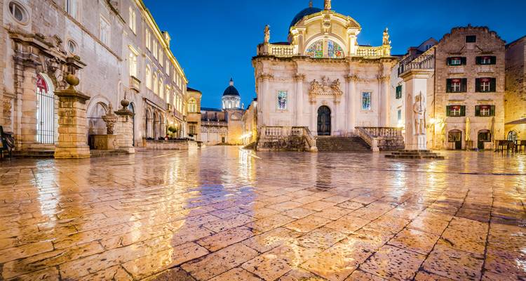 Rain-slicked Luža Square in Dubrovnik glistens at blue hour with St. Blaise Church illuminated against the night sky.