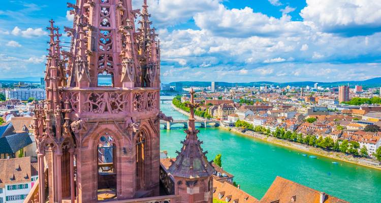 Le clocher gothique de la Cathédrale de Bâle surplombant le Rhin turquoise et les toits de la ville sous des nuages éclatants.