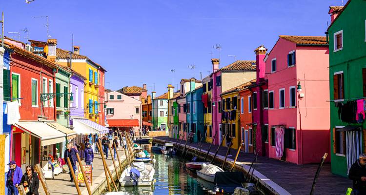 Les maisons aux couleurs vives bordent un canal à Burano avec des visiteurs se promenant devant les bateaux amarrés.