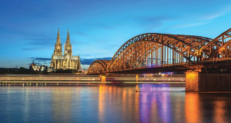 Der Kölner Dom und die illuminierte Hohenzollernbrücke spiegeln sich während der blauen Stunde im Rhein.