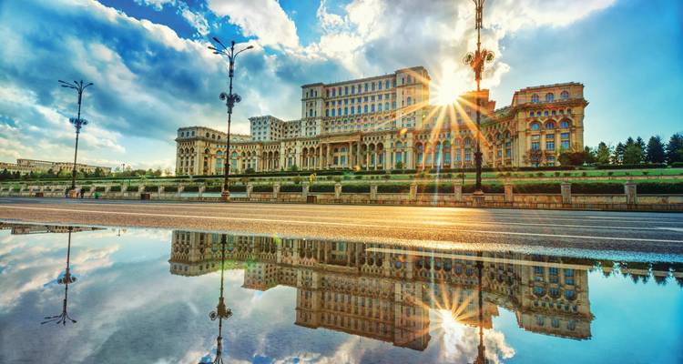 Le Palais du Parlement à Bucarest brille à l'heure dorée, se reflétant dans une flaque de pluie sous un ciel dramatique.