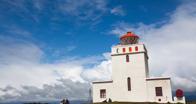 Le phare blanc avec sa lanterne d'un orange vif se dresse contre un ciel bleu profond tandis que les visiteurs se reposent sur la colline herbeuse en contrebas.
