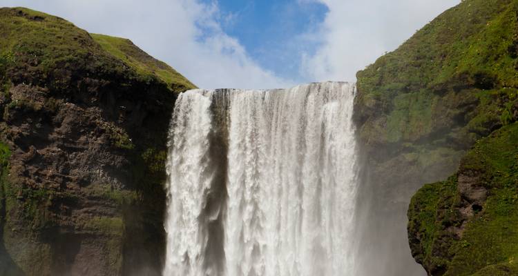 Gros plan de la puissante chute d'eau de Skógafoss se précipitant depuis des falaises couvertes de mousse sous des parcelles de ciel bleu.