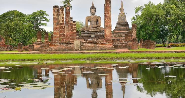 Ruhige Spiegelung eines sitzenden Buddha und Ziegelstupen in einem lotuserfüllten Teich im Sukhothai Historical Park.
