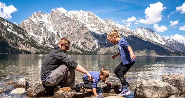 Papá e hijos juegan en la orilla rocosa del lago con montañas nevadas reflejadas en el agua cristalina.