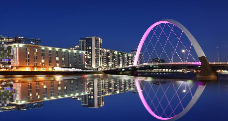 Puente de arco iluminado brillando en rosa y el perfil de la ciudad reflejándose perfectamente en un río tranquilo por la noche.
