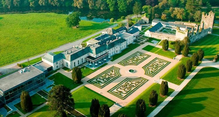 Vue aérienne d'un manoir élégant, de jardins formels et de ruines de château adjacentes, bordés par des pelouses verdoyantes et une rivière.