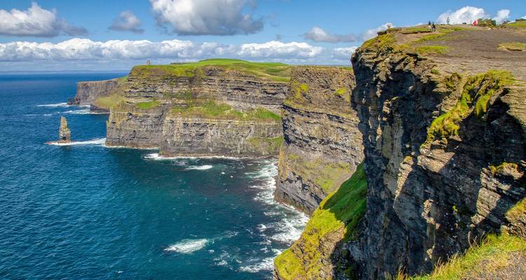 Des falaises majestueuses de l'Atlantique aux couches de roche abrupte plongent dans des eaux turquoise sous un ciel bleu avec des nuages cotonneux.
