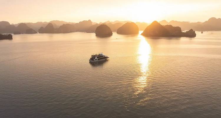 Vista aérea de un crucero deslizándose por las aguas doradas y tranquilas de la Bahía de Ha Long al atardecer con islas de piedra caliza recortadas contra el cielo.