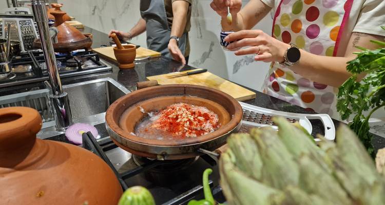Een deelnemer strooit kruiden in een kleiën tajine terwijl anderen groenten voorbereiden tijdens een Marokkaanse kookles.