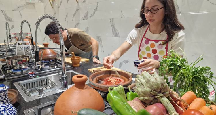 Vrouw in gestipte schort roert ingrediënten in een tajine omgeven door verse groenten en kruiden.