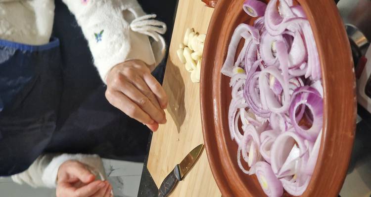 Close-up van handen die knoflook klaarmaken naast een tajine gevuld met gesneden rode uien op een snijplank.