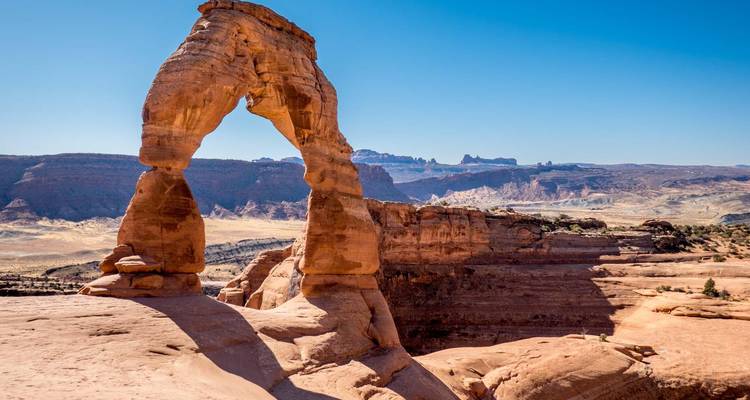 El icónico Arco Delicado se alza sobre las rocas lisas del desierto con mesetas lejanas bajo un cielo azul despejado
