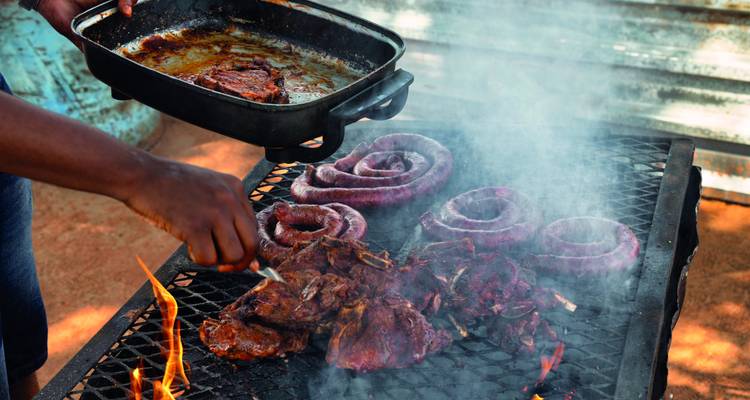 Küchenchef bei der Zubereitung von zischendem Fleisch und aufgerollter Wurst auf einem offenen Feuer eines südafrikanischen Braai-Grills.