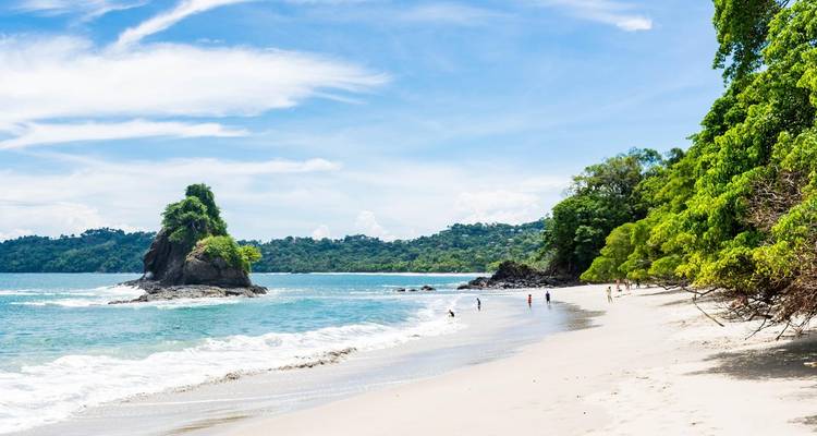 Pristine white-sand beach and turquoise water in Manuel Antonio National Park with small rocky islet and rainforest backdrop.