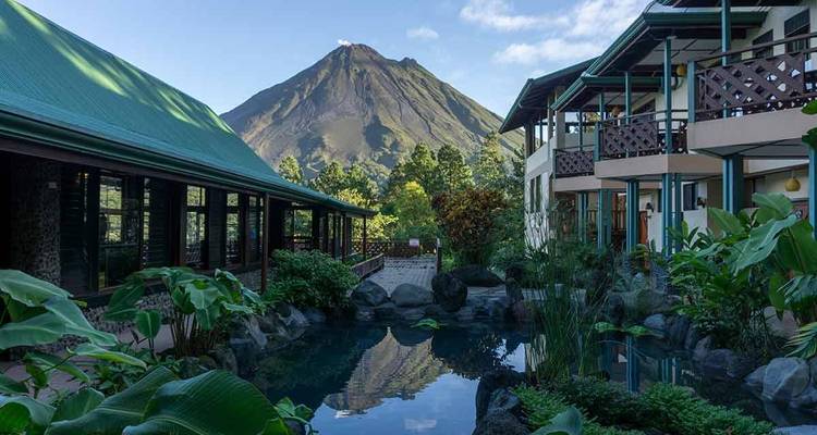 Resort courtyard with pool reflecting Arenal Volcano in the background under blue skies.