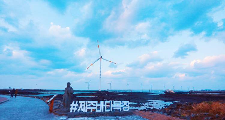 Windkraftanlagen drehen sich entlang der felsigen Küste von Jeju unter einem dramatischen Himmel und einem #Jeju-Schild.