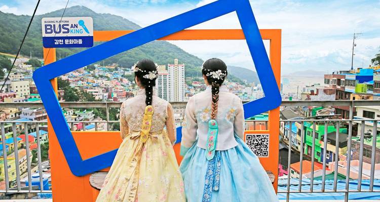 Zwei Frauen in farbenfrohen Hanbok blicken durch eine Fotorahmen-Installation über das Gamcheon Culture Village von Busan.