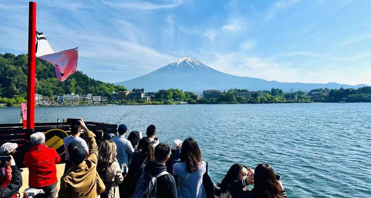 Turistas a bordo de un barco fotografiando el Monte Fuji nevado al otro lado de un lago tranquilo bajo un cielo azul despejado.