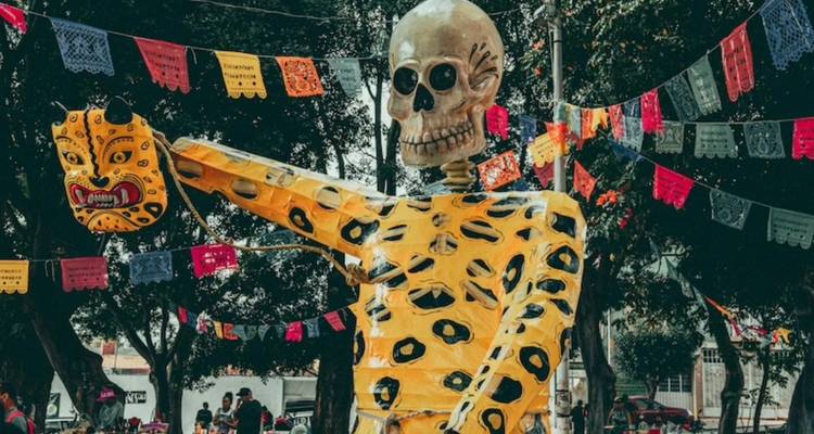 Estatua de esqueleto amarillo y alto con patrón de jaguar rodeada de banderitas de papel picado en una plaza festiva.