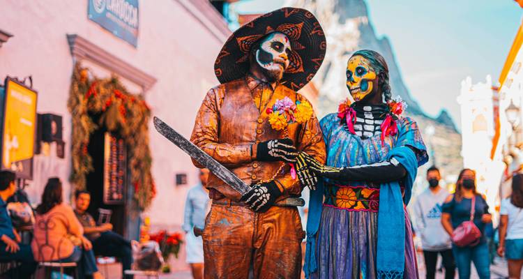 Couple de squelettes colorés du Jour des morts en costumes ornés posant avec des fleurs sur une rue mexicaine animée.