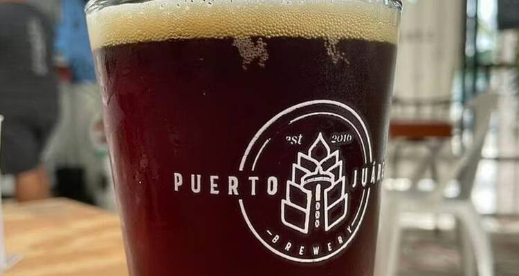 Nahaufnahme eines dunklen Craft-Beer-Glases mit dem Stempel des Puerto Juárez Brewery-Logos auf einem Holztisch.