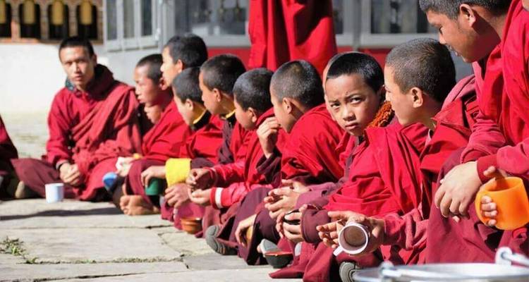 Rangée de jeunes moines en robes rouges assis à l'extérieur avec des tasses, écoutant attentivement.