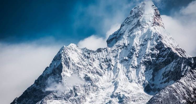 Pic himalayen dramatique couvert de neige s'élevant abruptement dans un ciel bleu chargé avec des nuages tourbillonnants.